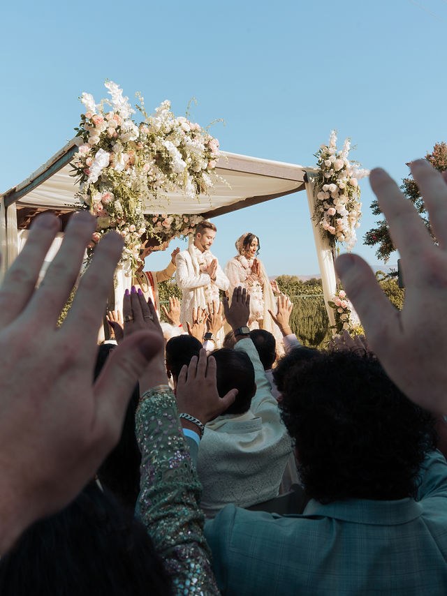 Guests raise their hands as Anika and Caden stand beneath the flower-framed canopy
