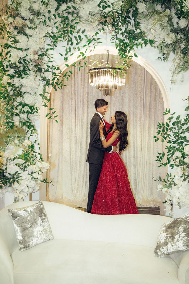 Karishma and Shil in evening attire beneath a chandelier and floral arch