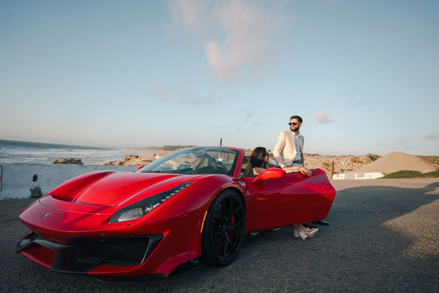 Paul and Gureet beside a red sports car on the coast
