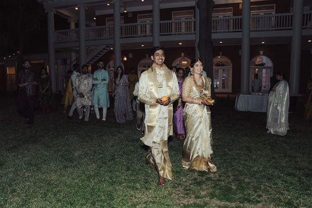 Rachana and Anoop walking toward their evening ceremony in ivory and gold